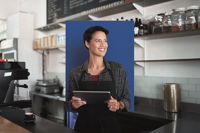 Eine junge Unternehmerin steht in ihrem Kaffeehaus. Sie lächelt und hält ein Tablet in den Händen.