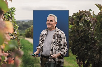 Ein Weinbauer steht in seinem Weingarten. Er hat eine Gartenschere und einen Eimer in der Hand.