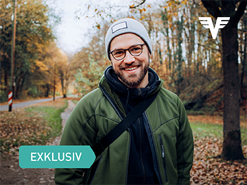 Ein Mann mit Mütze und Daunenjacke steht auf einem Spazierweg im Park und grinst in die Kamera.