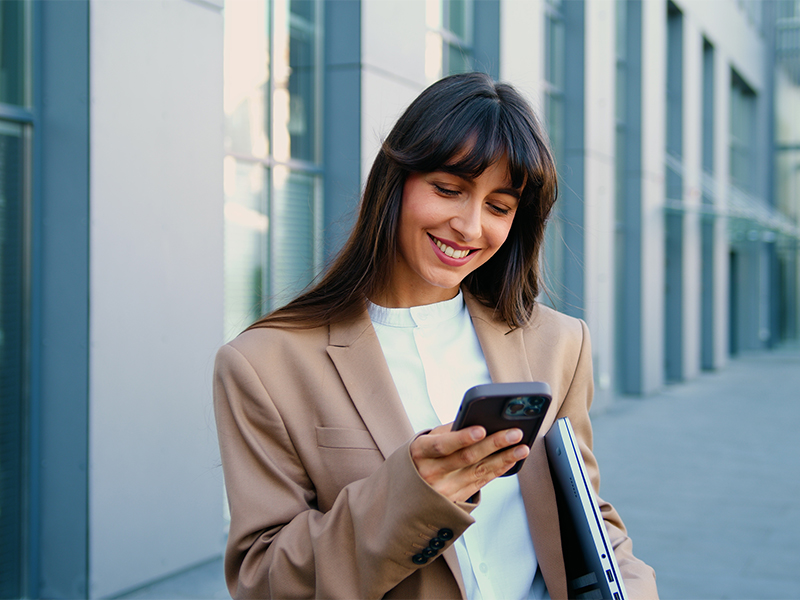 Eine Frau steht vor einem modernen Bürogebäude und blickt auf ein Smartphone, während sie einen Laptop unter dem Arm hält
