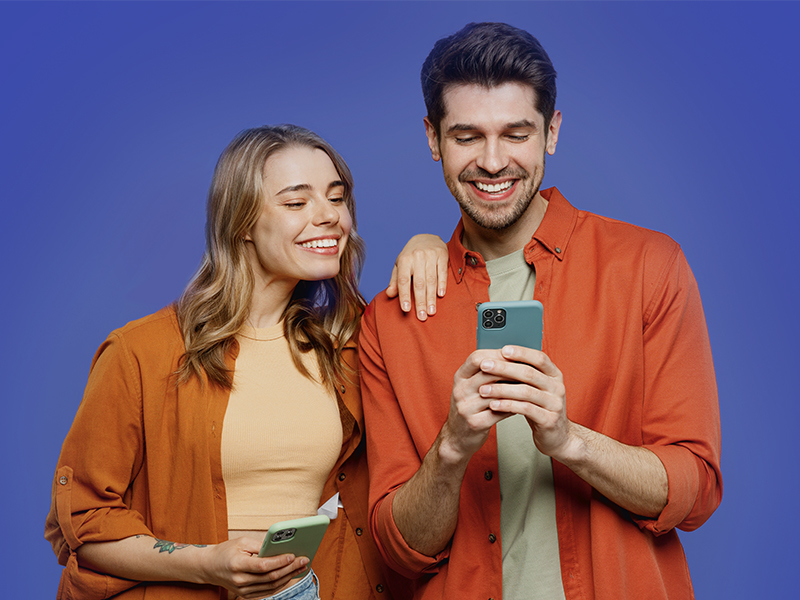 Eine junge Frau und ein junger Mann stehen vor einem violetten Hintergrund und betrachten gemeinsam Inhalte auf einem Smartphone.