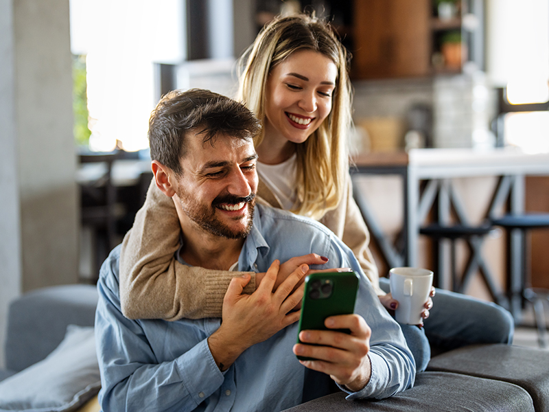 Eine Frau umarmt einen Mann während sie ihrer Wohnumgebung sind und sehen dabei gemeinsam auf ein Smartphone.