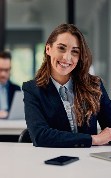 Eine Geschäftsfrau im Blazer sitzt lächelnd an einem Schreibttisch.