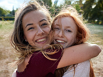 Zwei junge Frauen stehen auf eine Wiese und umarmen sich. Sie lachen beide in die Kamera.