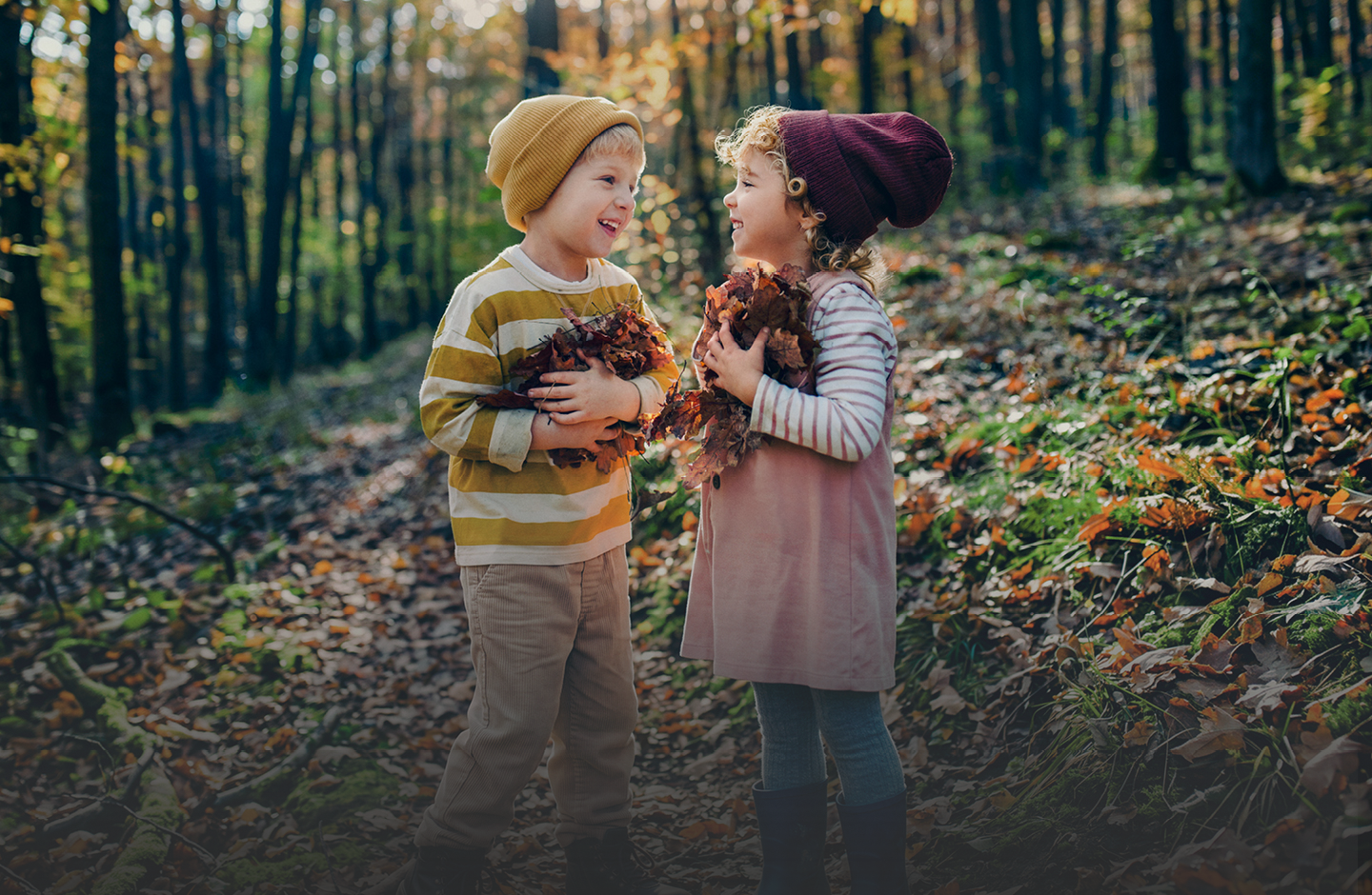 Zwei Kinder stehen in einem herbstlichen Wald. Beide haben trockene Blätter in den Armen.
