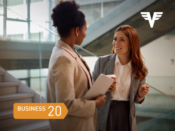 Zwei Frauen im Business-Outfit stehen im Stiegenhaus eines modernen Bürogebäudes und unterhalten sich.
