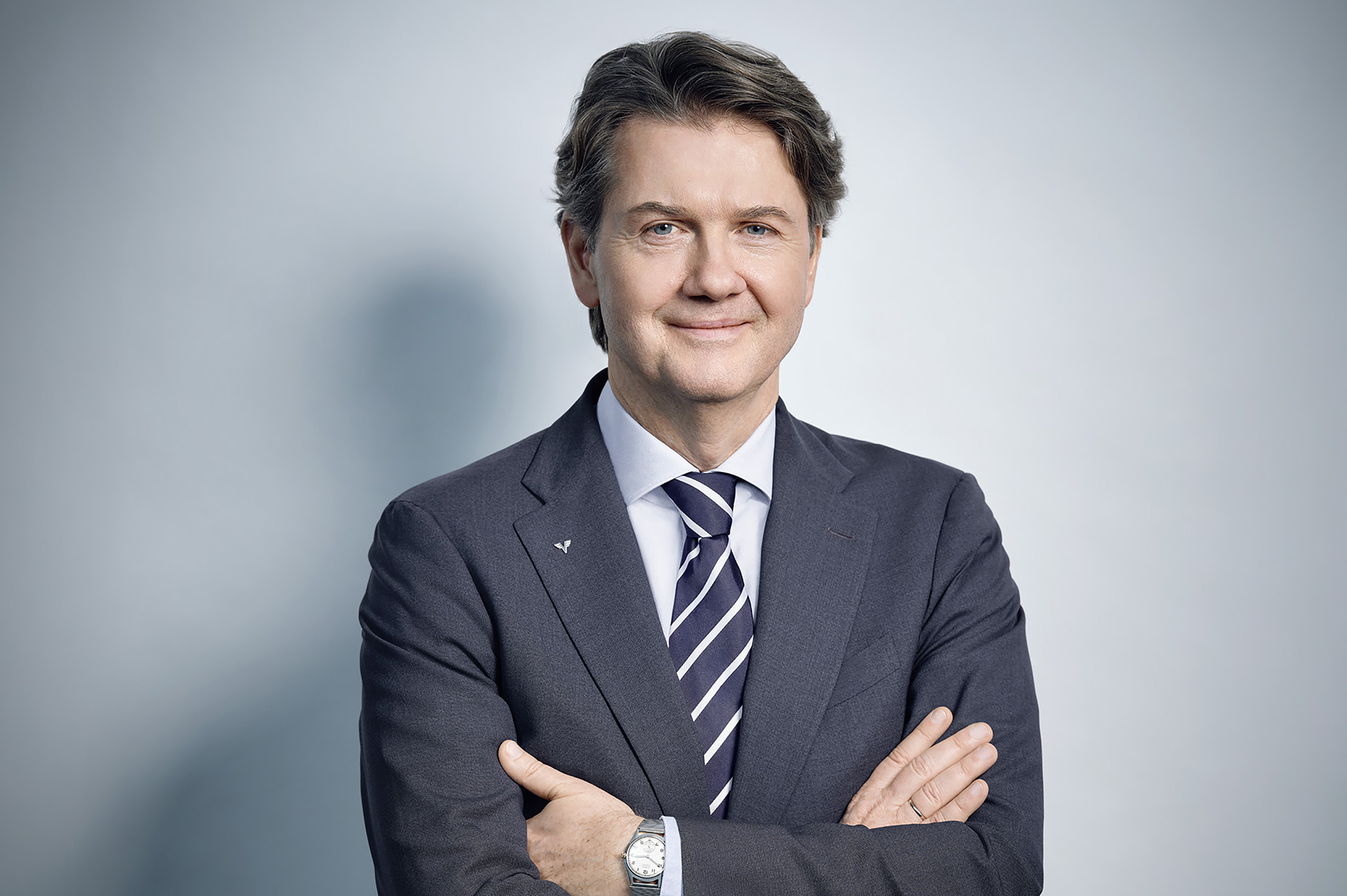 Gerald Fleischmann im Anzug mit verschränkten Armen auf hellblauem Hintergrund.