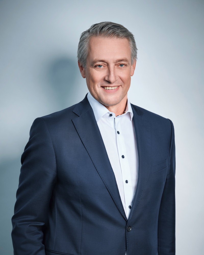 Rainer Borns im Anzug auf hellblauem Hintergrund.