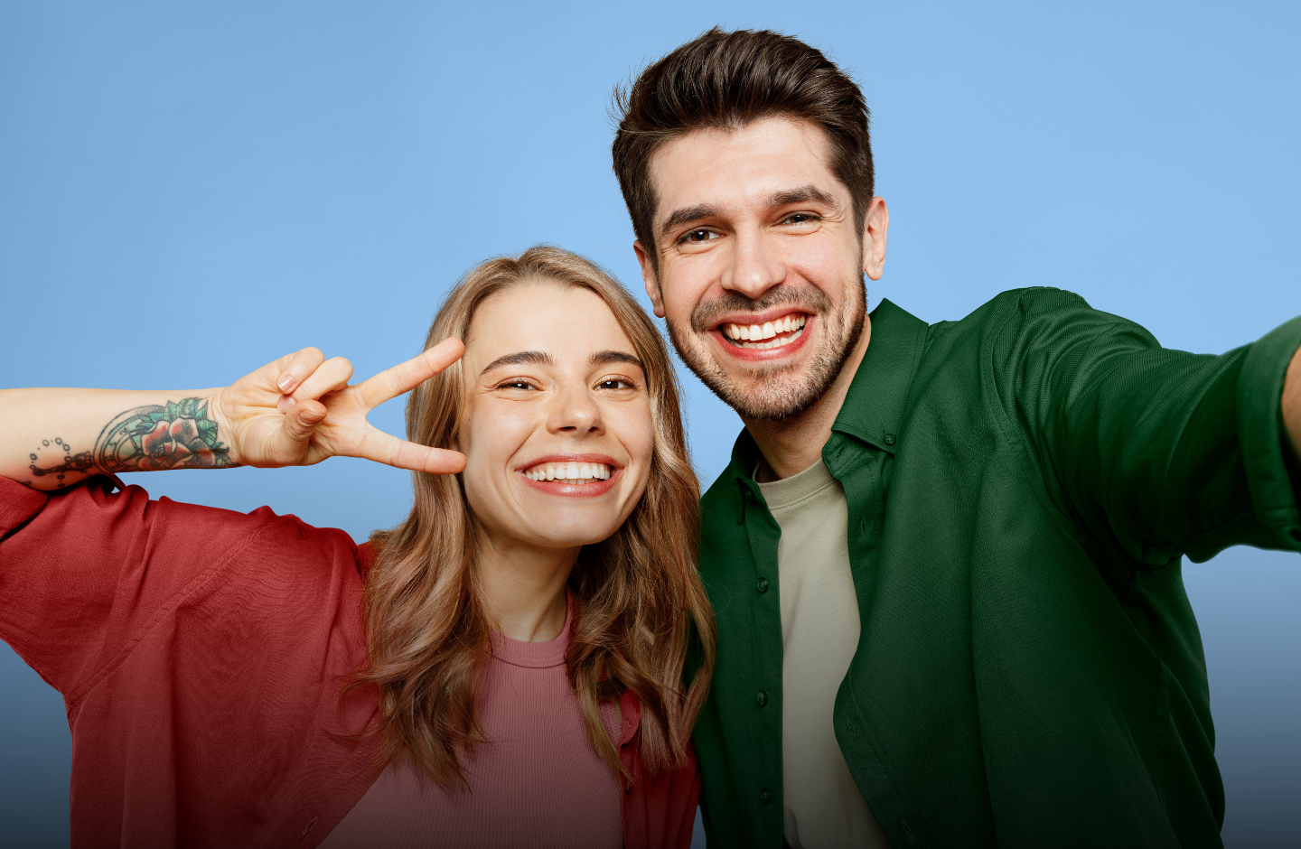 Eine junge Frau und ein junger Mann strahlen. Sie zeigt das Peace-Zeichen, während er die Kamera hält, als würden sie ein Selfie machen.