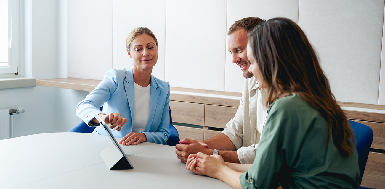 Eine Frau im Blazer berät ein Paar mithilfe eines Tablets in einem Büro.