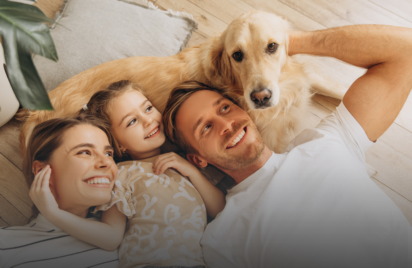 Eine Familie mit Hund liegt auf dem Boden. Vater, Mutter und Tochter lächeln den Hund an.