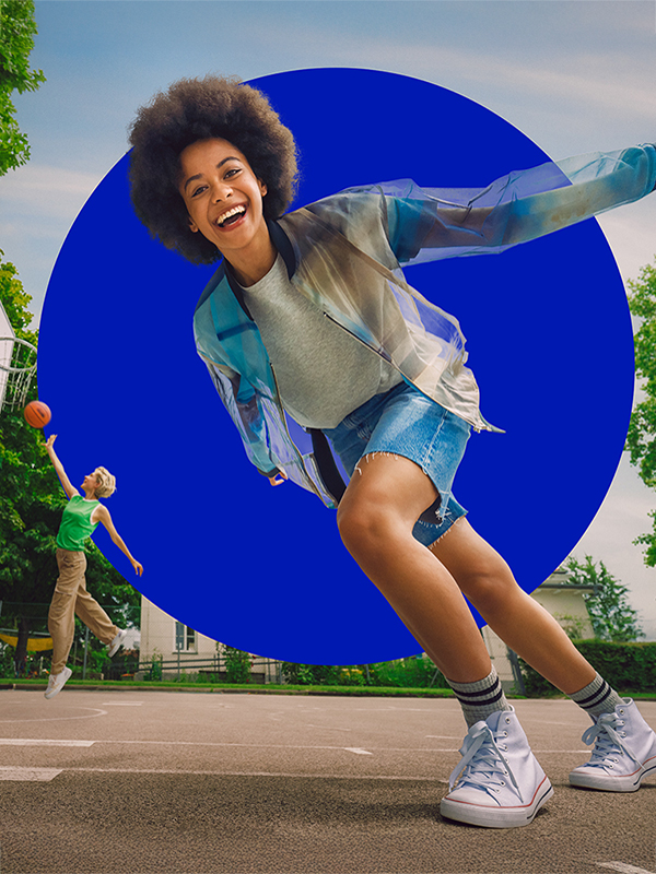 Zwei junge Frauen spielen Basketball. Eine der Frauen wirft im Springen einen Korb, die andere läuft an der Kamera vorbei und lacht.