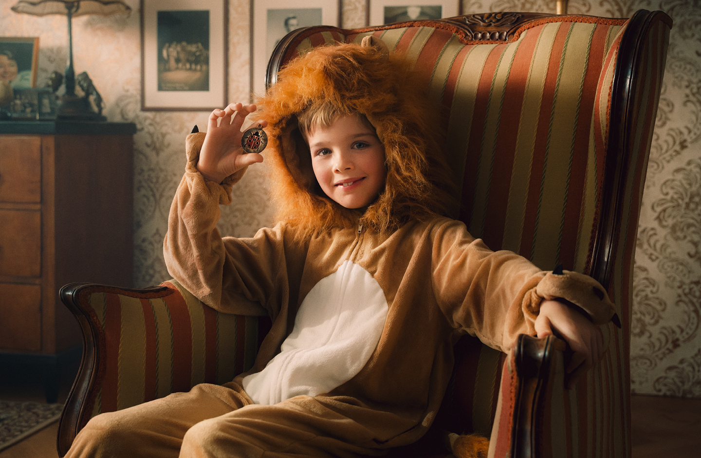 Ein kleiner Junge sitzt in einem Löwenkostüm auf einem altmodischen Sessel und lächelt. Er hält einen Talisman in der Hand.