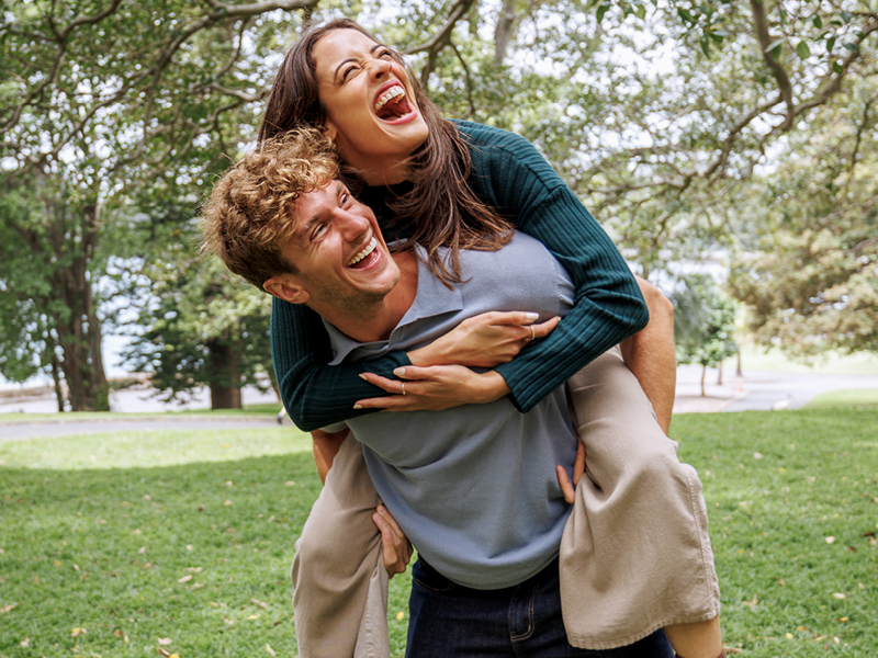 Ein Mann und eine Frau sind in einem Park an einem See. Er hat sie huckepack. Sie lachen beide ausgelassen.