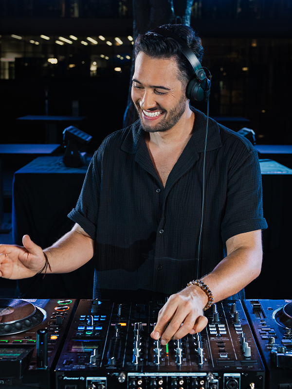 Ein DJ in einem schwarzen Hemd steht for seinen Turntables und legt Musik auf. Er hat Kopfhörer auf und lacht.