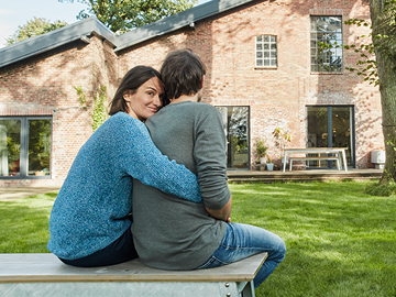 Ein Mann und eine Frau sitzen mit dem Rücken zur Kamera vor einem Haus. Die Frau umarmt den Mann von hinten. Sie blickt über die Schulter in die Kamera.