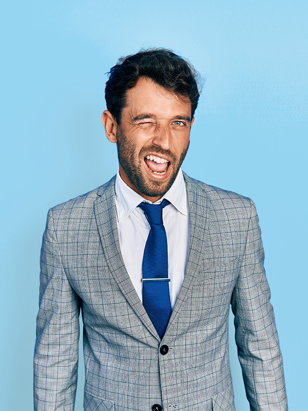 Ein Mann im grauen Anzug steht vor hellblauem Hintergrund und wzinkert in die Kamera.