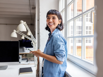 Eine junge Frau steht vor einer Fensterfront im Büro und hält lachend ihr Handy in der Hand.