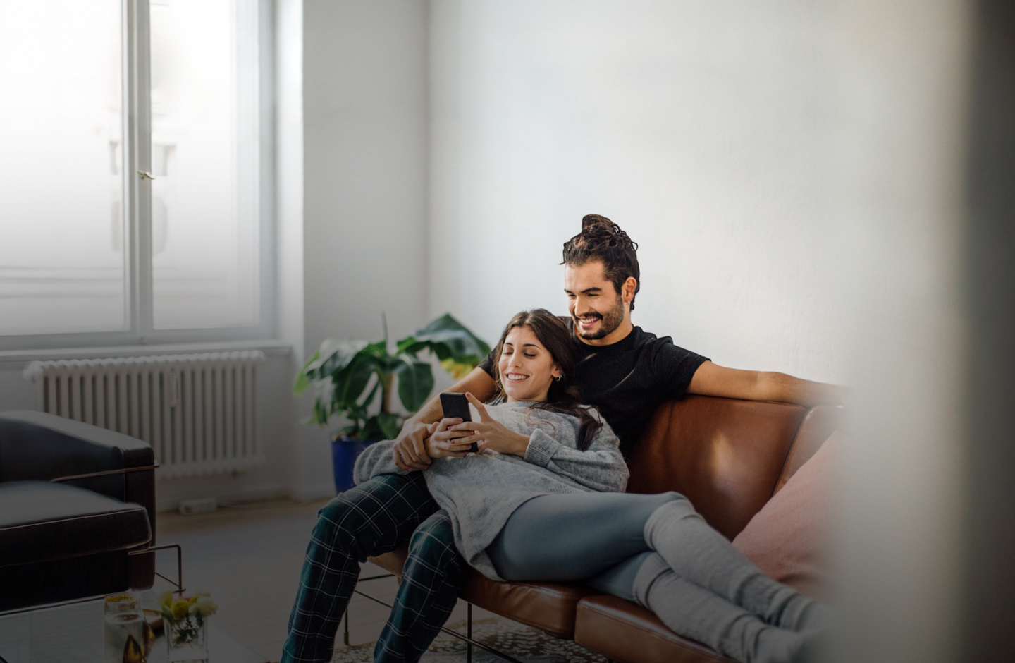 Eine Frau liegt in den Armen eines Mannes auf einer Couch. Gemeinsam blicken sie auf ein Smartphone.