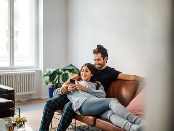 Eine Frau liegt in den Armen eines Mannes auf einer Couch. Gemeinsam blicken Sie auf ein Handy.