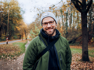 Ein Mann mit Mütze und Daunenjacke steht auf einem Spazierweg im Park und grinst in die Kamera.